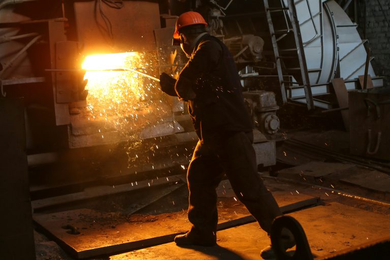 A worker in protective gear welds metal in an industrial plant with sparks flying.